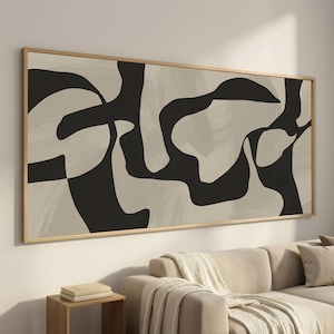Op de afbeelding: Abstracte kunstprint met een beige achtergrond en gedurfde zwarte vormen. Het kunstwerk is ingelijst in een natuurlijk hout, wat een moderne, minimalistische uitstraling geeft. Het canvas wordt boven een bank gepresenteerd.
