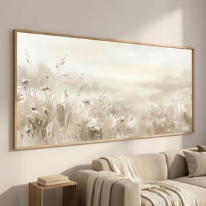 May include: A framed landscape painting featuring a field of tall grasses and wildflowers in muted beige and white tones. The artwork has a soft, blurred effect, creating a serene and natural scene. The frame is a light wood color.