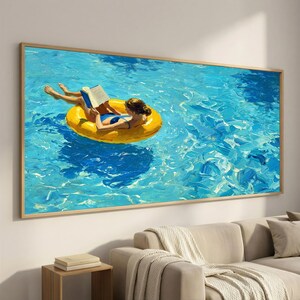 May include: A rectangular wall art print featuring an oil painting of a person in a blue swimsuit floating on a bright yellow circular pool float in vibrant blue water while reading an open book. The artwork is framed in a thin light wood frame.