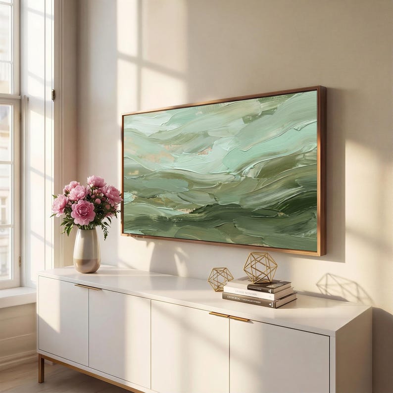 May include: An abstract painting with a wooden frame, featuring shades of green and white brushstrokes. The artwork hangs above a white cabinet with gold handles. A vase of pink flowers and decorative gold geometric shapes are also present.