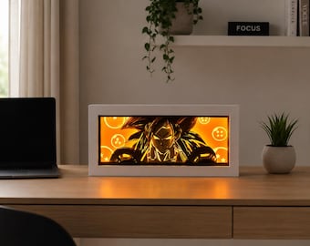 Könnte beinhalten: Ein rechteckiger weißer Leuchtkasten mit einem mehrschichtigen Papierschnitt-Design eines Anime-Charakters mit stacheligen Haaren. Das interne warme orangefarbene Licht beleuchtet die Silhouette und mehrere kreisförmige Drachenball-Symbole.