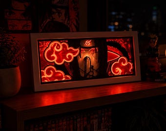 Könnte beinhalten: Eine rechteckige Leuchtbox mit einem mehrschichtigen Papierschnitt-Design des Akatsuki-Wolkensymbols und einer zentralen Charakterfigur. Das Kunstwerk wird von innen durch ein leuchtend rotes Licht beleuchtet und ist in einem weißen Rahmen gefasst.