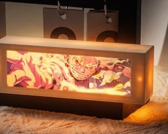 Könnte beinhalten: Ein rechteckiges Nachtlicht mit einem Cartoon-Bild, das in einem weißen Rahmen eingefasst ist. Das Bild ist durch ein rechteckiges Fenster sichtbar und zeigt eine Figur mit entschlossenem Ausdruck. Im Hintergrund ist ein Kalender zu sehen. Das Licht strahlt warm.