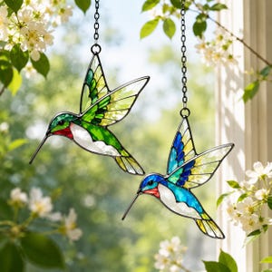 May include: Two stained glass hummingbird ornaments hanging from black metal chains. One bird features green, yellow, and white glass with a red throat patch. The second bird features blue, yellow, and white glass with a red throat patch.