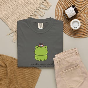 Puede incluir: Camiseta gris oscuro Comfort Colors con una rana de dibujos animados que lleva un sombrero pequeño. La rana es verde con vientre blanco y contornos negros. El texto "I am the executive of this dysfunction" está impreso debajo.