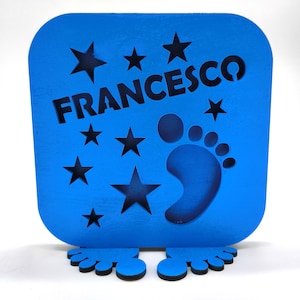 Puede incluir: Un letrero de madera azul con el nombre "FRANCESCO" recortado, estrellas y un diseño de huella. El letrero tiene una base en forma de pie. El diseño está pintado en un color azul vibrante.