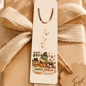 May include: A rectangular white bookmark featuring a cartoon frog wearing a witch hat, surrounded by autumn-themed items and the text Sweet Spells & Cozy Treats, tied to a brown gift box with a beige ribbon and a For You tag.