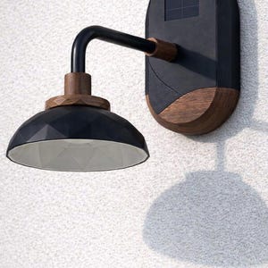 Op de afbeelding: Zwarte en houten buitenwandlamp. De lamp heeft een geometrische kap, een gebogen arm en een zonnepaneel. Gemonteerd op een witte, getextureerde muur, werpt het een schaduw.
