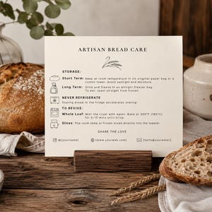 May include: A cream-colored card titled ARTISAN BREAD CARE with black text and line icons. It provides instructions for storage, reviving, and freezing bread, including temperatures like 350°F. The card is held upright by a dark wooden block stand.