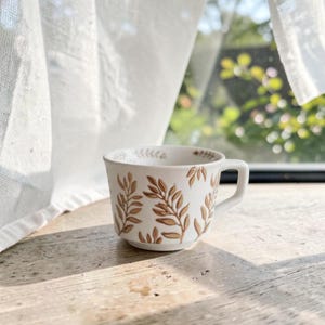 Può includere: Una tazza in ceramica bianca con un motivo ripetuto di rami frondosi dipinti a mano in marrone. La tazza presenta un bordo largo e svasato, un manico robusto sul lato destro e una base affusolata con un dettagliato design botanico.