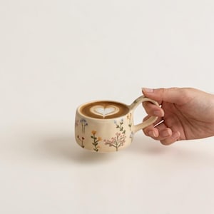 Könnte beinhalten: Ein cremefarbener Keramikbecher mit einem zarten, handgemalten Muster aus kleinen, bunten Wildblumen in Rosa-, Blau- und Gelbtönen. Der Becher ist mit einem Kaffeegetränk gefüllt, das mit einem herzförmigen Milchschaummuster verziert ist.