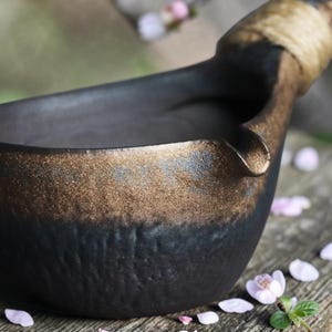 Puede incluir: Un cuenco de cerámica negra hecho a mano con una superficie texturizada y un borde dorado. El asa está envuelta con una cuerda de color natural. El cuenco está diseñado con un pico para verter.