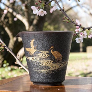Puede incluir: Una jarra de té de cerámica gris oscuro con un pico, con una ilustración dorada de dos grullas y un diseño de río que fluye. La jarra está sobre una superficie de madera, con un fondo borroso de flores de cerezo.