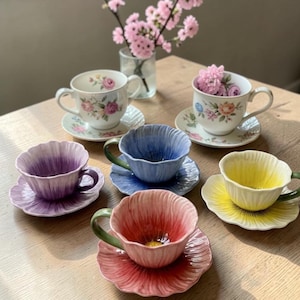 Puede incluir: Un conjunto de seis tazas y platillos. Dos tazas tienen diseños florales, mientras que las otras cuatro son de colores lisos: morado, azul, rojo y amarillo. Cada juego está sobre una mesa de madera, con un jarrón de flores rosas al fondo.