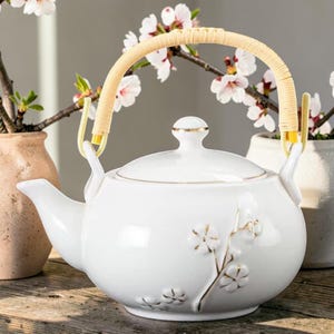 Puede incluir: Una tetera de cerámica blanca con asa trenzada y detalles dorados. La tetera presenta un diseño floral y una tapa con ribete dorado. El asa es de un material tejido de color claro. La tetera está sobre una superficie de madera.