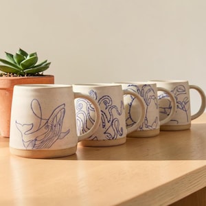 Könnte beinhalten: Ein Set aus vier cremefarbenen Keramikbechern mit gesprenkelter Textur, die jeweils eine durchgehende blaue Strichzeichnung eines anderen Meerestieres zeigen, darunter ein Wal, ein Oktopus, eine Krabbe und ein Hummer, mit einem unglasierten Boden.