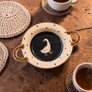 Può includere: Una ciotola rotonda in ceramica con due manici laterali, caratterizzata da un bordo color crema con pois neri e un centro scuro e maculato decorato con l'illustrazione di un'oca bianca. La ciotola è circondata da diversi sottobicchieri rotondi in maglia beige.