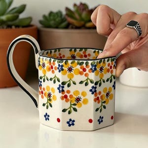 May include: A white ceramic mug with a hexagonal shape and a black handle. The mug is decorated with a floral pattern in yellow, orange, and blue. The rim is outlined in black.