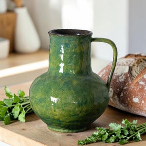 May include: A green ceramic jug with a handle. The jug has a textured surface and a dark brown rim. Fresh coriander and a loaf of bread are on a wooden surface in the background. The jug is a decorative kitchen item.