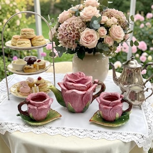 Op de afbeelding: Een keramieken theeservies in de vorm van roze rozen met groene blaadjes als schoteltjes, inclusief een theepot en twee kopjes. Een metalen etagère met scones en gebakjes staat naast een zilveren theepot en een boeket roze rozen.