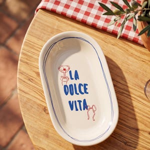 May include: An oval, off-white serving platter with a blue outline and the words "LA DOLCE VITA" in blue. The platter has a red and blue cocktail illustration and a red bow illustration. The platter is on a wooden table.