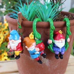 Könnte beinhalten: Drei farbenfrohe Gartenzwergfiguren, die am Rand eines Terrakottatopfes hängen. Die Zwerge tragen rote, blaue und violette Outfits mit weißen Bärten und Zipfelmützen. Ein Zwerg hält einen Korb mit Beeren.