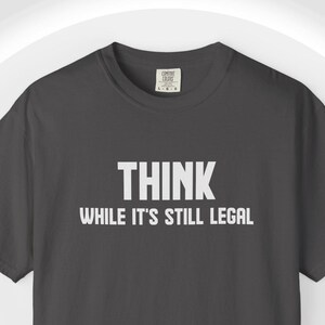 May include: A dark grey t-shirt featuring the white text "THINK WHILE IT'S STILL LEGAL". The short-sleeved shirt has a crew neck. A simple, casual style of garment.
