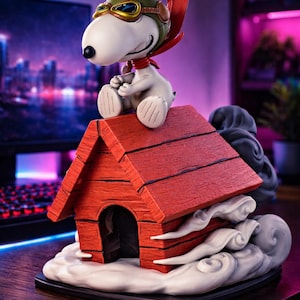 Może przedstawiać: Figurka Snoopy'ego jako asa lotnictwa z I wojny światowej, siedząca na swoim czerwonym domku dla psów. Snoopy ma na sobie zielony hełm lotniczy, gogle i czerwony szalik. Domek dla psów stoi na podstawie z chmurą, z panoramą miasta w tle.