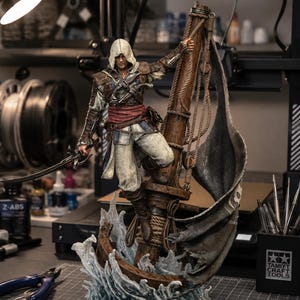 May include: A detailed figurine of a hooded pirate character standing on a wooden ship mast, holding a sword in one hand and a rope in the other. The figure wears a white hood, brown leather armor, and a red sash, set against a base of sculpted blue waves.