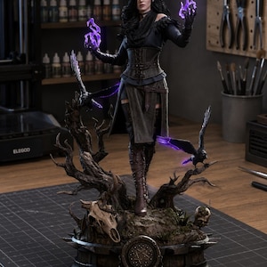 May include: A detailed figurine of a dark-haired sorceress wearing a black corset and boots, standing on a base with a skull and two ravens. The figure holds glowing purple magical energy in both hands, with a pentagram emblem on the front base.