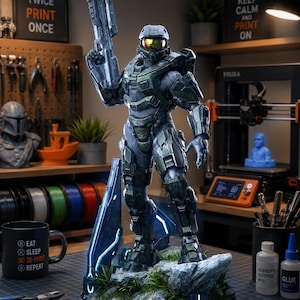 Puede incluir: Una figura detallada de Master Chief sobre una base rocosa con follaje verde y detalles de luz azul. El personaje viste una armadura verde oliva, una visera dorada y sostiene un rifle futurista en un entorno de taller.