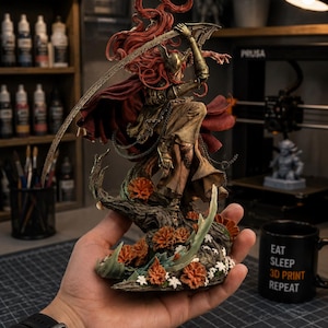 Puede incluir: Una figura pintada a mano de un guerrero con largo cabello rojo, armadura dorada y capa roja oscura, sosteniendo una hoja curva. La figura se encuentra sobre una base decorada con flores naranjas y follaje verde, mostrando una pose dinámica.
