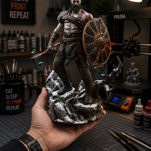 Può includere: Una statuetta dettagliata di un guerriero barbuto con un segno rosso sul corpo, che impugna un'ascia metallica e uno scudo circolare. Il personaggio poggia su una base rocciosa innevata con un ramo nodoso, caratterizzato da texture complesse.