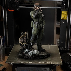 Könnte beinhalten: Detaillierte Figur einer Figur in grüner Militäruniform, die eine Zigarre hält, steht auf einem Sockel mit einer Schlange und Blumen. Die Figur befindet sich auf einem 3D-Drucker. Der Bildschirm des Druckers zeigt "PRUSA i3 MK3S+ READY."