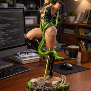 Puede incluir: Estatua detallada de una guerrera en una pose dinámica, vestida con un atuendo verde y negro. La figura sostiene un bastón y se encuentra sobre una base con calaveras y cadenas. La estatua está sobre un escritorio de madera con una computadora y otros artículos de oficina.