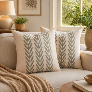 May include: Two square throw pillows with a cream-colored background and a repeating pattern of gray leaves. The pillows are placed on a beige sofa, with a tan throw blanket draped over the arm. A lamp and a small potted plant are visible in the background.