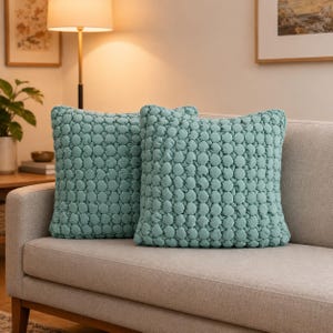 May include: Two square, pale teal cushions with a textured, quilted design. They are placed on a light grey sofa, creating a cosy atmosphere. The cushions add colour and texture to the neutral-toned living space. Each cushion measures approximately 40 cm x 40 cm.