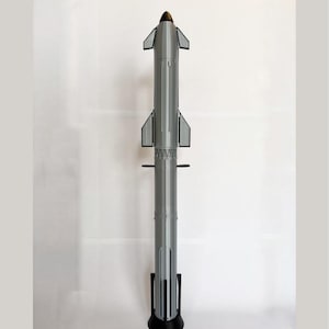 Pode incluir: Um modelo de foguete cinza e preto, em pé. O foguete tem um nariz preto pontudo com uma ponta dourada, barbatanas cinzentas e uma base preta. O foguete é uma réplica detalhada.