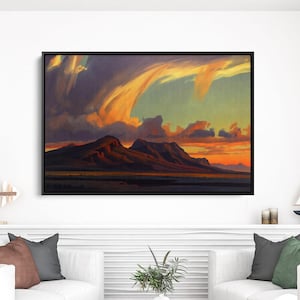 May include: A framed landscape painting depicts a mountain range under a dramatic sky. The artwork features warm colors, with orange and yellow hues dominating the sky and a dark, silhouetted mountain range. The painting is displayed above two white sofas.