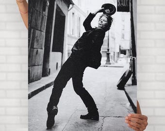 May include: Black and white print of a person in a black outfit throwing a hat in the air on a city street. The person is leaning back with one leg extended. The image is held by a person.