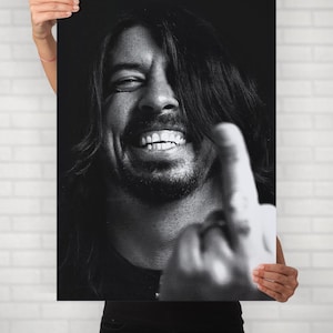 May include: Black and white print of a smiling person with long hair, giving the middle finger. The image is held up by a person in front of a white brick wall. The person in the image is smiling with their teeth showing.