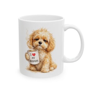 May include: White ceramic mug featuring a watercolor illustration of a fluffy, light brown dog holding a coffee mug. The mug the dog is holding says "I ❤️ MY HOOMAN" with a red heart. The dog has a sad expression and steam is rising from the coffee.