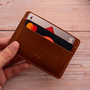 Pode incluir: Um porta-cartões de couro castanho com costuras visíveis ao longo das bordas e as letras YC gravadas na frente. O porta-cartões contém dois cartões de crédito, um dos quais apresenta um fundo preto com um logótipo circular vermelho e laranja.