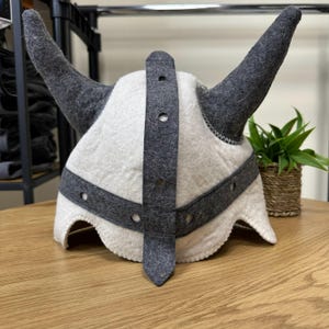 Puede incluir: Un casco de lana afieltrada en color blanco roto con dos grandes cuernos de color gris oscuro a los lados. El casco incluye una banda gris oscuro alrededor de la base y una tira vertical en el centro, ambas con aberturas circulares.