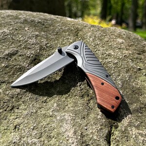 May include: A partially open pocket knife with a gray blade and a handle featuring a combination of gray metal and wood. The knife is resting on a textured gray rock, with a blurred green background.