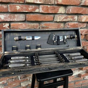 Könnte beinhalten: Ein dunkles Holz-Messerset mit einer Vielzahl von Messern und Zubehör. Das Set enthält ein großes Hackmesser, ein kleines Messer und mehrere Spieße, die alle ordentlich in einer Holzkiste angeordnet sind. Die Kiste ist geöffnet.