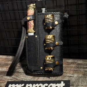 May include: A black leather-wrapped flask with a crocodile-skin texture, featuring a side holster holding a knife with a wolf-head handle and three gold-toned metal shot cups secured by leather straps with brass-coloured snap buttons.