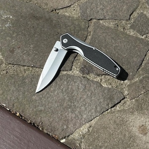 Könnte beinhalten: Ein silbernes und schwarzes Klappmesser mit teilweise geöffneter Klinge. Die Klinge ist silberfarben und der Griff ist schwarz mit einem strukturierten Muster. Das Messer liegt auf einer Steinoberfläche.