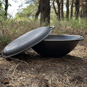 May include: A black cast iron cooking pot with a lid, resting on a bed of pine needles. The pot has a rounded shape with a silver rim and a handle on the lid. The lid is slightly ajar, revealing the interior.