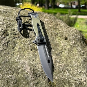 Könnte beinhalten: Ein Klappmesser mit schwarzer Klinge und grünem Griff. Das Messer hat eine gezahnte Kante und einen schwarzen Taschenclip. Am Ende des Griffs ist eine schwarze Kordel befestigt. Das Messer liegt auf einem großen, strukturierten Felsen.